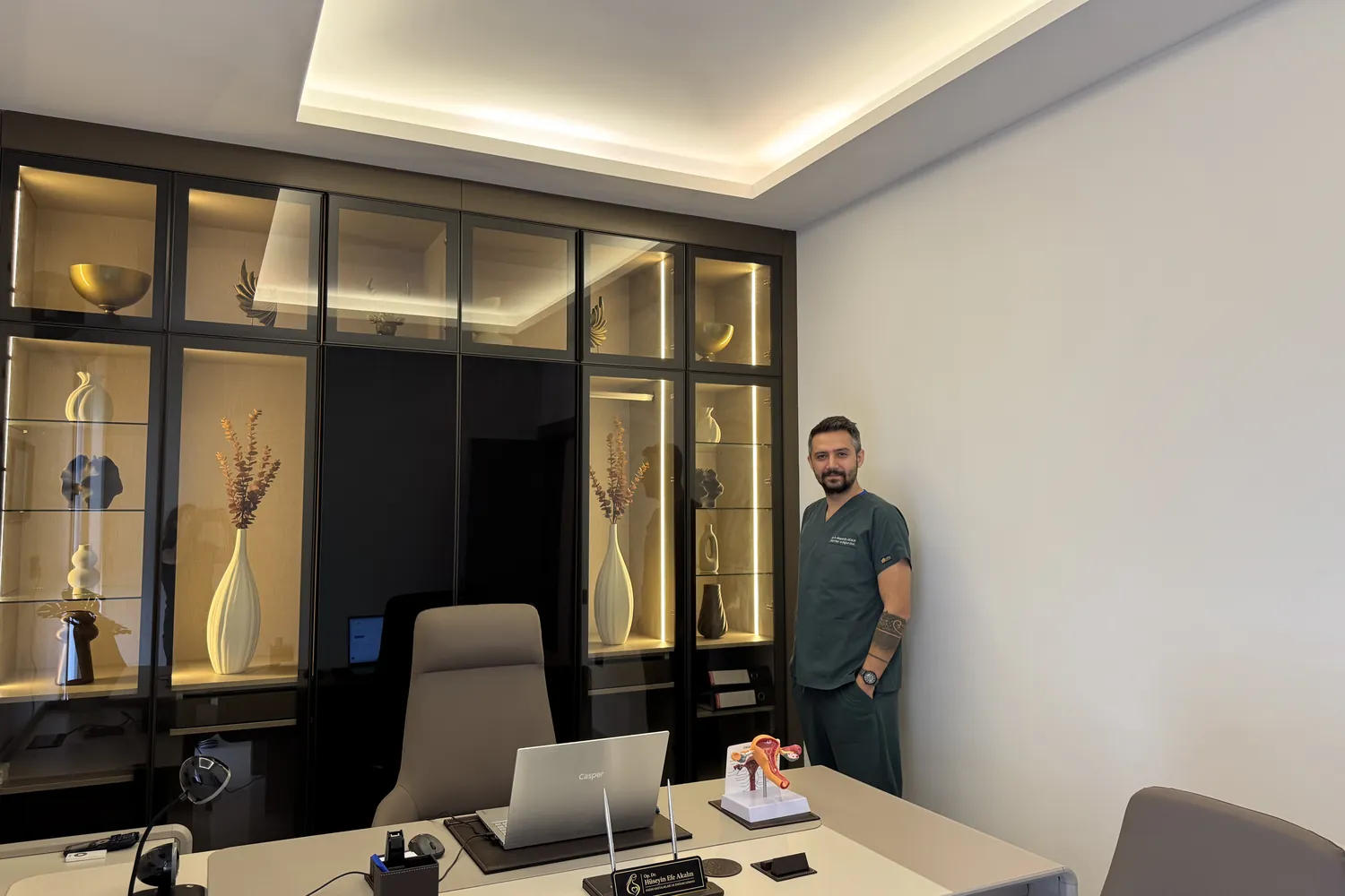 Op. Dr. Hüseyin Efe Akalın in the consultation room with lit display cabinets behind